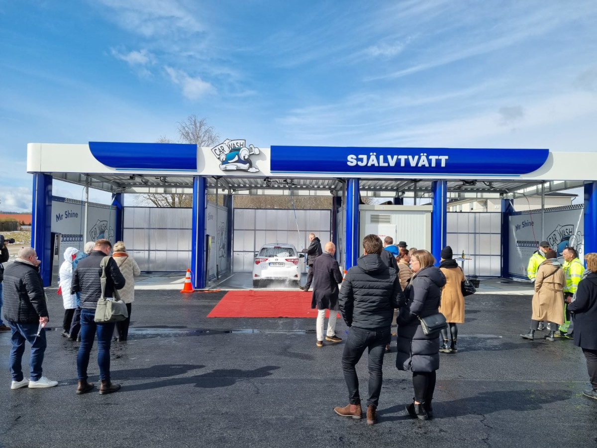 Automatyczna myjnia samochodowa z białym samochodem w środku i ludźmi na zewnątrz.